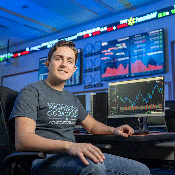 An Indiana State business student with short brown hair and wearing a gray Scott College of Business t-shirt sits next to a computer in the Scott College of Business virtual trading floor. The computer is displaying graphical stock data. On the wall behind, large monitors display more detailed stock data. Above that, an electronic stock ticker shows current trading values – Walmart is the one visible company name. Soft, electric blue light illuminates the scene.